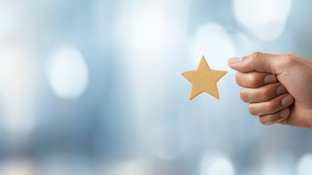 Close-up de uma m&atilde;o masculina segurando uma pequena estrela dourada entre os dedos. O fundo &eacute; um azul claro com luzes "bokeh" (c&iacute;rculos de luz desfocados), sugerindo um ambiente de excel&ecirc;ncia, avalia&ccedil;&atilde;o positiva ou sucesso em conquistas de marca.