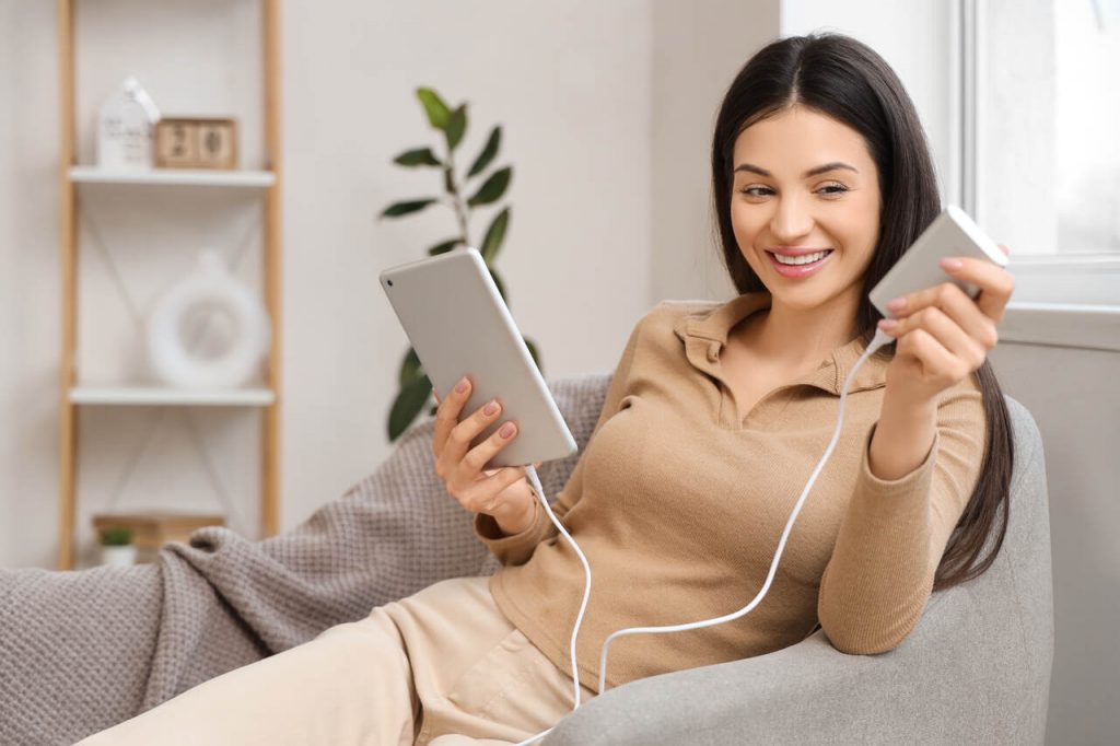 Uma mulher sorridente sentada em uma poltrona cinza, utilizando um carregador port&aacute;til prata para carregar seu tablet. Ela segura o tablet em uma m&atilde;o e o power bank na outra, demonstrando conveni&ecirc;ncia e conforto. O ambiente &eacute; uma sala de estar clara e moderna com uma estante ao fundo.
