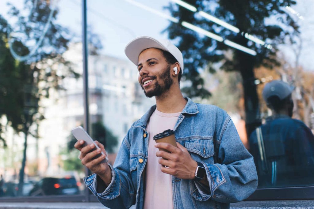 Um homem jovem em um ambiente urbano ensolarado, usando fones de ouvido sem fio e um smartwatch. Ele segura um smartphone e um copo de caf&eacute;, vestindo uma jaqueta jeans e bon&eacute; branco. A imagem destaca o uso de acess&oacute;rios tecnol&oacute;gicos no dia a dia e o estilo de vida moderno.