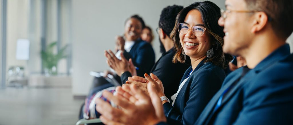 Profissionais sentados em um auditório corporativo aplaudindo, com uma mulher sorridente olhando para a câmera durante um evento empresarial.