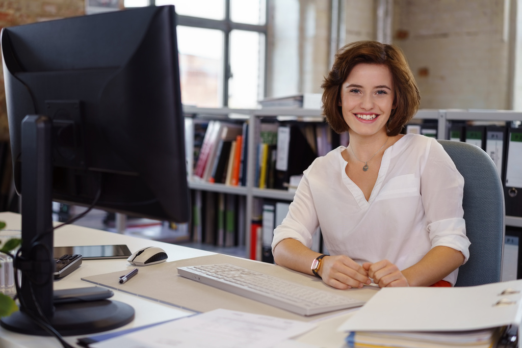 A mesma mulher da imagem anterior est&aacute; sentada em sua mesa de trabalho, sorrindo diretamente para a c&acirc;mera. &Agrave; sua frente, h&aacute; um monitor de computador, teclado e pap&eacute;is organizados. O ambiente de escrit&oacute;rio ao fundo &eacute; amplo, com janelas grandes e prateleiras repletas de pastas de arquivo.