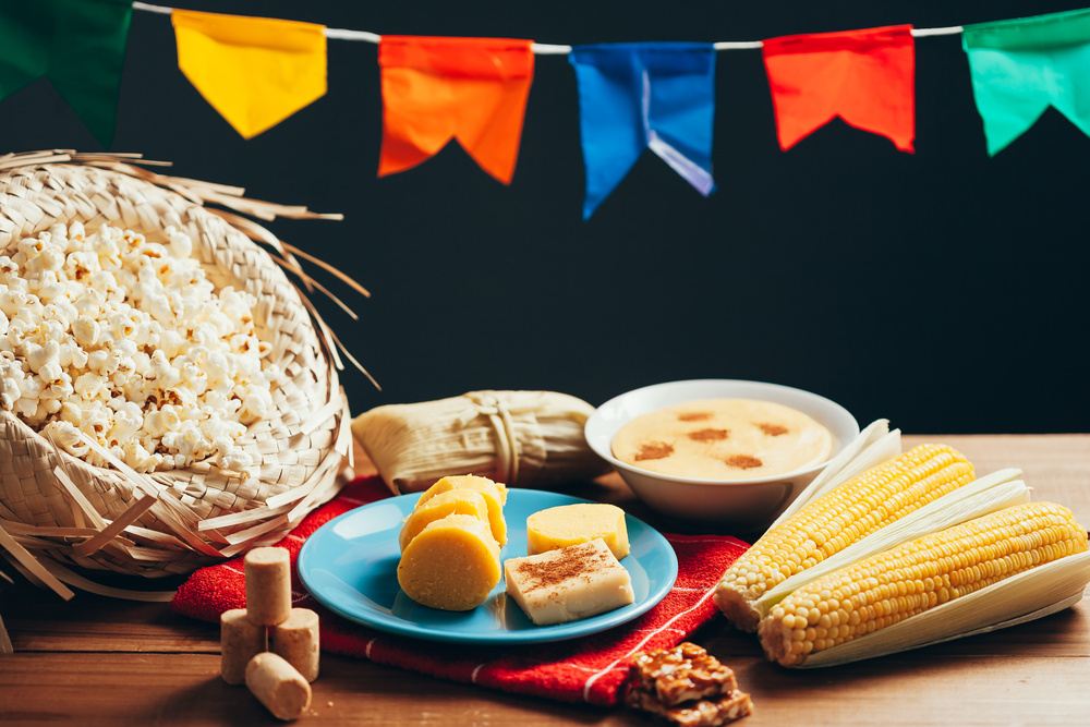 Alimentos juninos organizados sobre mesa decorada, com destaque para milho, doces e pipoca, inspira&ccedil;&atilde;o de lembrancinhas para as festas juninas.