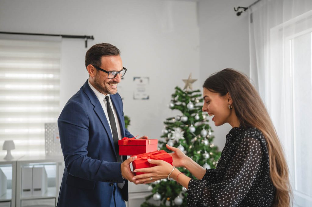 Homem sorridente em um terno azul marinho entrega duas caixas de presentes vermelhas com la&ccedil;os de fita para uma mulher em um escrit&oacute;rio decorado. Ao fundo, v&ecirc;-se uma &aacute;rvore de Natal enfeitada, sugerindo uma celebra&ccedil;&atilde;o de fim de ano ou premia&ccedil;&atilde;o corporativa de alto n&iacute;vel.