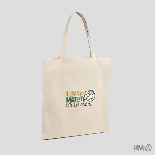 Uma sacola de tecido ecol&oacute;gica (ecobag) na cor bege claro, vista de &acirc;ngulo lateral. No centro, h&aacute; uma estampa colorida com o nome "Hakuna Matata Brindes" e o desenho estilizado de um le&atilde;o. O fundo &eacute; cinza claro e minimalista.