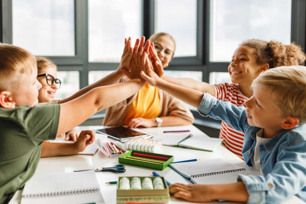 Grupo de quatro crian&ccedil;as, meninos e meninas de diversas etnias, unindo as m&atilde;os ao centro em um gesto de "high five" coletivo sobre uma mesa escolar. Ao fundo, uma professora sorri observando a integra&ccedil;&atilde;o. Sobre a mesa, h&aacute; materiais escolares como l&aacute;pis de cor, cadernos e estojos.
