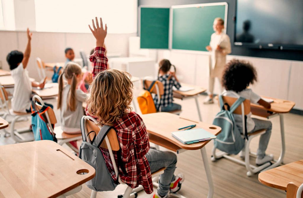 Vista do fundo de uma sala de aula iluminada onde v&aacute;rios alunos est&atilde;o sentados em suas carteiras. Um aluno em destaque, vestindo uma camisa xadrez vermelha, est&aacute; com a m&atilde;o levantada para participar da aula. Ao fundo, a professora est&aacute; em p&eacute; pr&oacute;xima ao quadro verde e a uma tela digital.