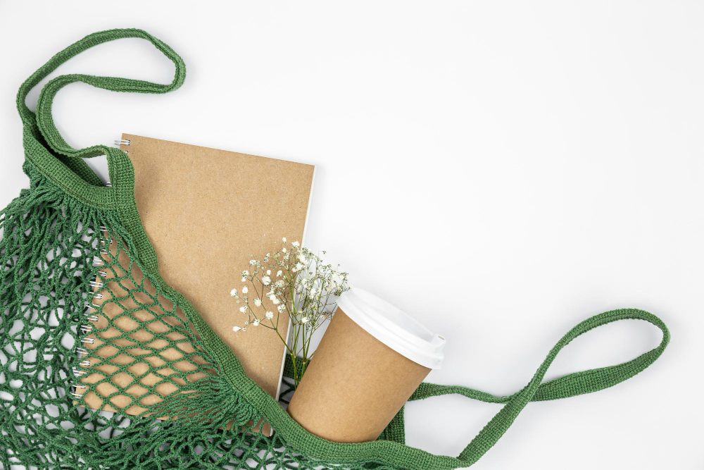 Sacola ecológica verde com itens sustentáveis como copo reutilizável, papel kraft e flores secas.