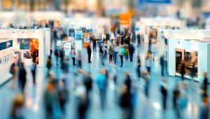 Vista aérea de um grande saguão de feira de eventos ou convenção corporativa. A imagem utiliza um efeito de desfoque de movimento, mostrando diversas pessoas circulando entre estandes brancos e modernos. Transmite a sensação de um ambiente dinâmico, movimentado e cheio de interações.