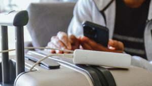 Close-up de um carregador portátil (power bank) branco sobre uma mala de viagem bege. Um cabo de carregamento conecta o dispositivo a um smartphone, que é segurado por uma pessoa ao fundo