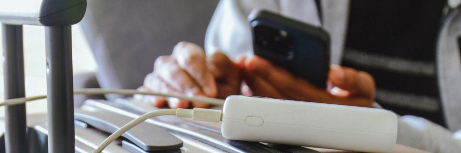 Close-up de um carregador portátil (power bank) branco sobre uma mala de viagem bege. Um cabo de carregamento conecta o dispositivo a um smartphone, que é segurado por uma pessoa ao fundo