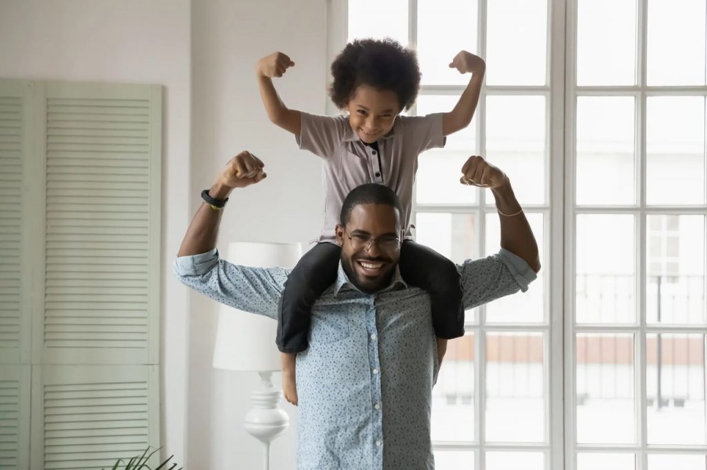 Pai com filhos nos ombros, ambos fazendo uma pose com os braços flexionados para cima, demonstrando força