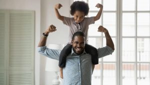 Pai com filhos nos ombros, ambos fazendo uma pose com os braços flexionados para cima, demonstrando força