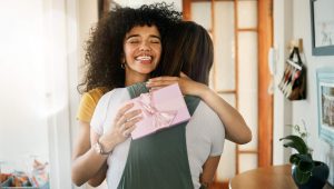 Uma cena calorosa e espontânea de duas amigas se abraçando. A mulher em destaque tem cabelos escuros e cacheados e sorri com felicidade, de olhos fechados. Ela segura uma pequena caixa de presente rosa claro, decorada com um laço de fita de cetim, encostada nas costas da outra pessoa durante o abraço. O ambiente sugere a entrada de uma casa iluminada.