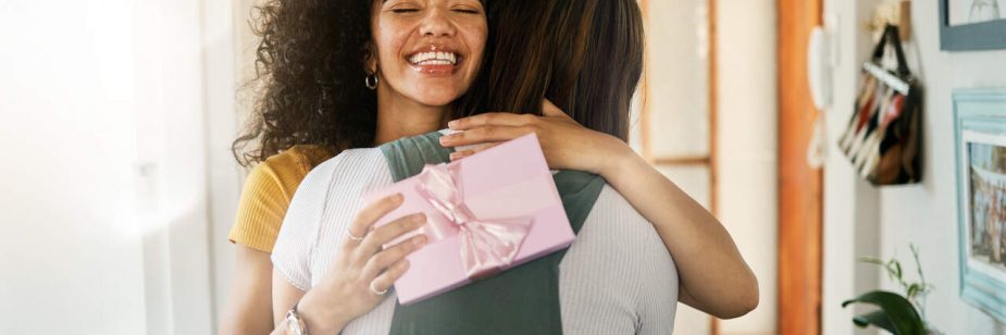 Uma cena calorosa e espontânea de duas amigas se abraçando. A mulher em destaque tem cabelos escuros e cacheados e sorri com felicidade, de olhos fechados. Ela segura uma pequena caixa de presente rosa claro, decorada com um laço de fita de cetim, encostada nas costas da outra pessoa durante o abraço. O ambiente sugere a entrada de uma casa iluminada.