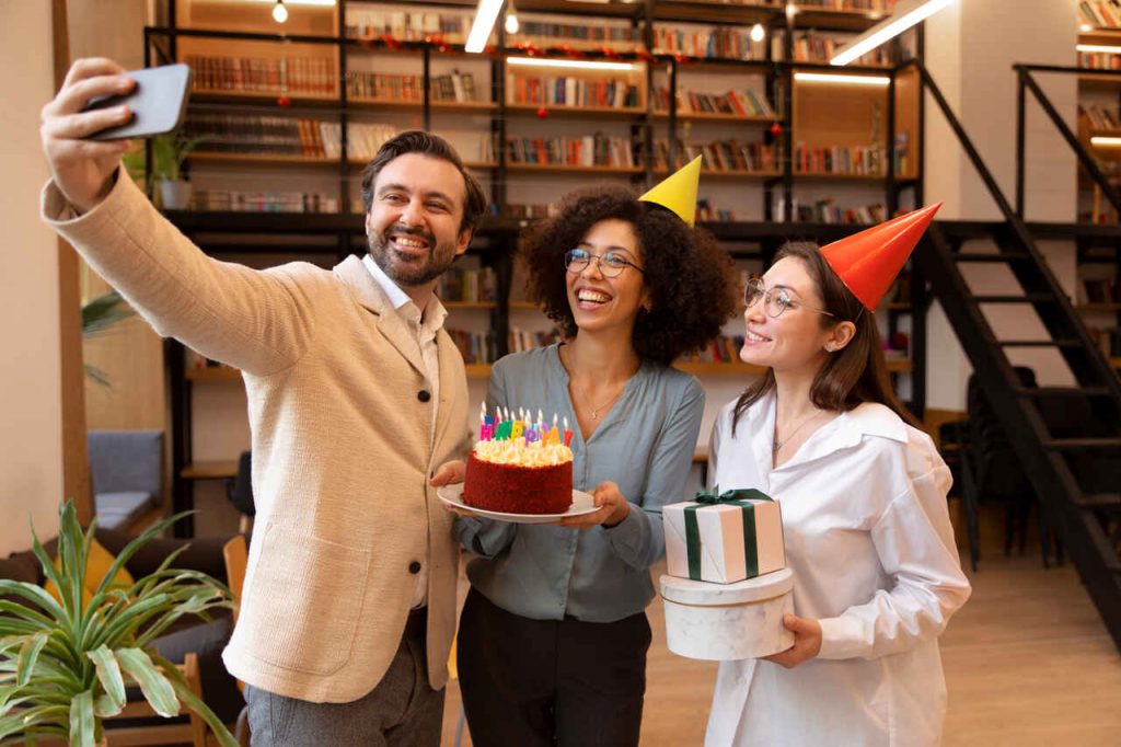 Três colegas de trabalho sorridentes celebram um aniversário em um ambiente de escritório moderno com estantes de livros ao fundo. À esquerda, um homem tira uma selfie do grupo; no centro, uma mulher segura um bolo com velas coloridas acesas; à direita, outra mulher usando um chapéu de festa laranja segura duas caixas de presente empilhadas.