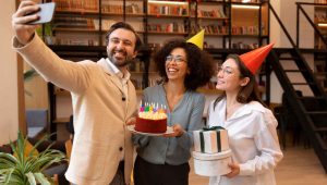 Três colegas de trabalho sorridentes celebram um aniversário em um ambiente de escritório moderno com estantes de livros ao fundo. À esquerda, um homem tira uma selfie do grupo; no centro, uma mulher segura um bolo com velas coloridas acesas; à direita, outra mulher usando um chapéu de festa laranja segura duas caixas de presente empilhadas.