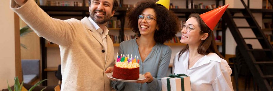 Três colegas de trabalho sorridentes celebram um aniversário em um ambiente de escritório moderno com estantes de livros ao fundo. À esquerda, um homem tira uma selfie do grupo; no centro, uma mulher segura um bolo com velas coloridas acesas; à direita, outra mulher usando um chapéu de festa laranja segura duas caixas de presente empilhadas.