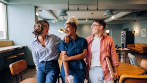 Três colegas conversando e sorrindo em um ambiente de trabalho moderno, transmitindo união e clima leve no escritório.