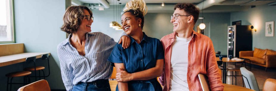 Três colegas conversando e sorrindo em um ambiente de trabalho moderno, transmitindo união e clima leve no escritório.