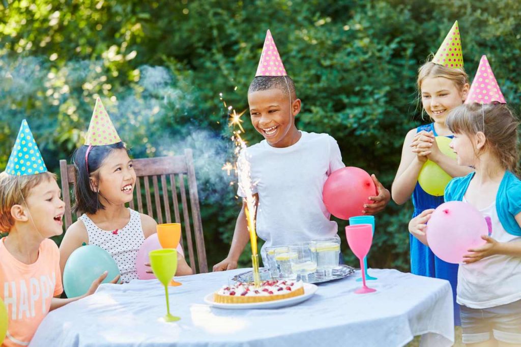 Festa infantil com crianças sorrindo de chapéu, segurando balões, com vela acessa sobre torta na mesa e taças de plástico ao redor
