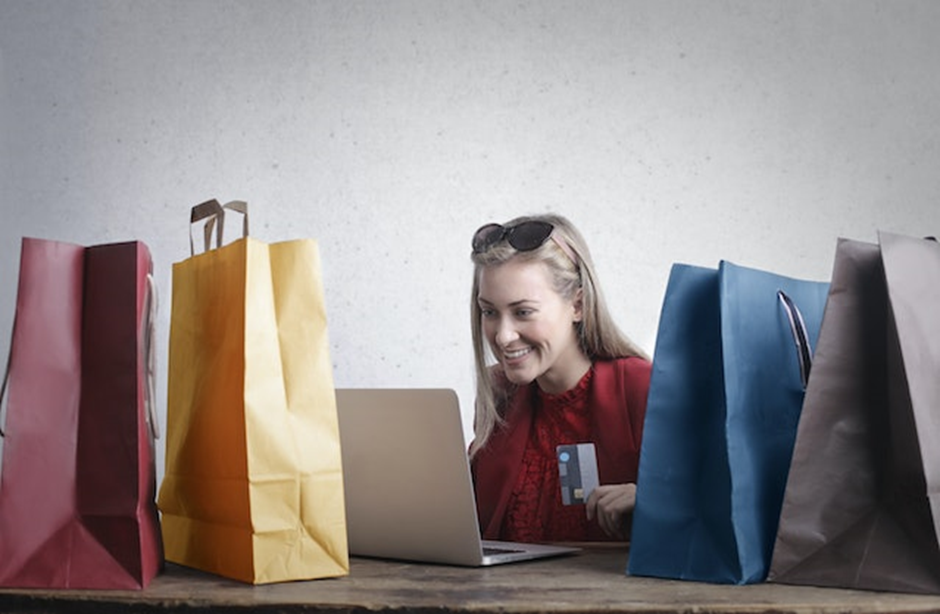 Mulher segurando cartão de crédito sorrindo, olhando para tela do computador, com quatros sacolas de compras coloridas ao redor, simbolizando cliente e fortalecimento de marca