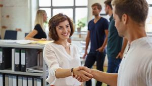 Uma mulher jovem de cabelos curtos castanhos e blusa branca sorri alegremente enquanto aperta a mão de um colega em um escritório moderno. Ao fundo, outras pessoas conversam em um ambiente de trabalho iluminado e casual, com estantes de arquivos organizadas à esquerda.