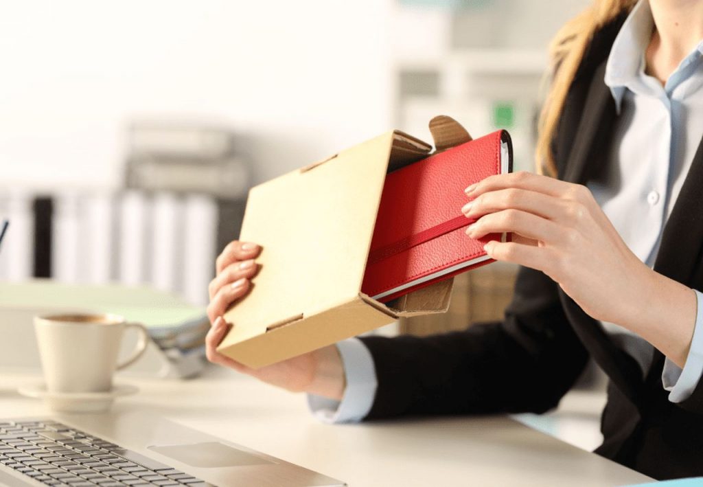 Pessoa em escritório tirando um caderno tipo moleskine de dentro de uma embalagem de papelão, simbolizando ideias de mimos para funcionários