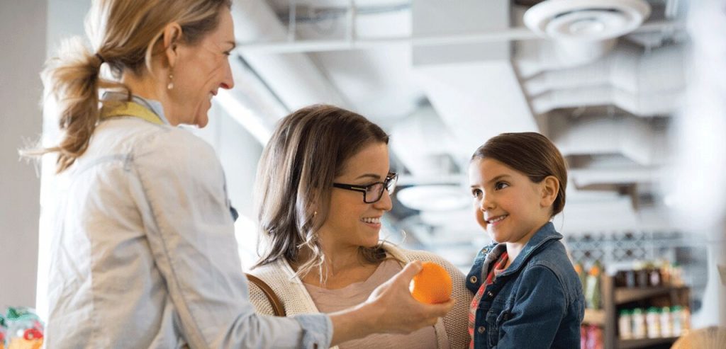 Simbolizando a semana do cliente, atendente entrega uma fruta laranja para uma criança atenta sorridente que está no colo da mãe que também sorri