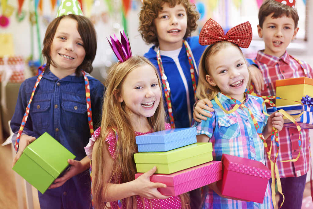 Crianças com enfeites com chapeuzinho, lacinho e fitas decorativas, celebrando segurando caixas de presente que simbolizam lembrancinha para alunos