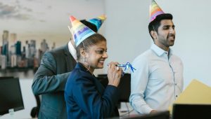 Pessoas no escritório usando chapéus de festa e assoprando adereços em celebração de aniversário de empresa.