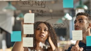 Dois profissionais, uma mulher negra com cabelos cacheados e um homem jovem, olham atentamente para uma parede de vidro transparente em um escritório moderno. Eles estão organizando post-its coloridos e escreveram a palavra "Branding" no vidro com um marcador. A cena foca na expressão de concentração e planejamento estratégico, ilustrando um processo de brainstorming e estruturação de marca.