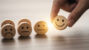 Mão escolhendo um bloco de madeira com rosto sorridente entre rostos tristes e neutros, simbolizando a jornada de satisfação do cliente