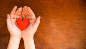 Mãos em formato de concha segurando um pequeno coração vermelho sobre uma mesa de madeira.