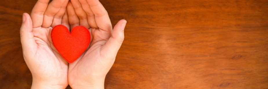 Mãos em formato de concha segurando um pequeno coração vermelho sobre uma mesa de madeira.