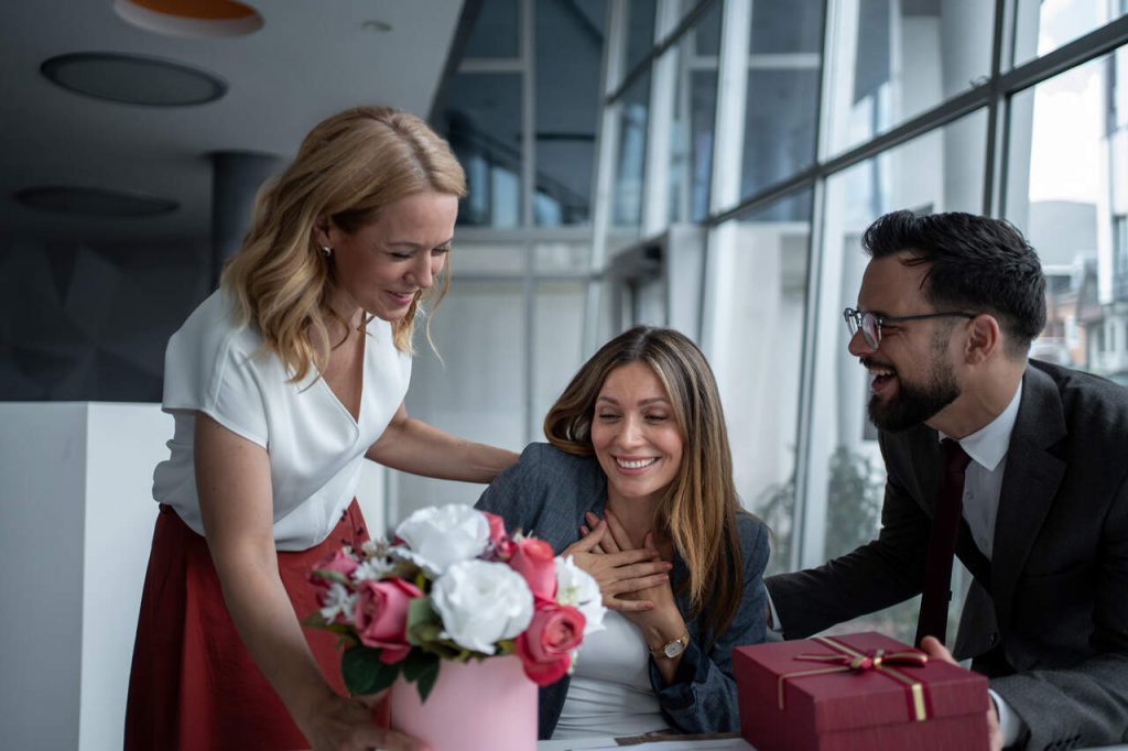 Três colegas de trabalho em um ambiente de escritório moderno celebram um momento especial. Uma mulher ao centro parece emocionada e surpresa ao receber um arranjo de flores e uma caixa de presente elegante de seus colegas. A cena transmite gratidão e o impacto emocional de brindes de luxo personalizados.