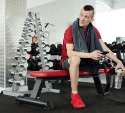 Pessoa sentada em banco de academia segura uma squeeze vermelha, destaque entre os equipamentos ao fundo. Ideal como brindes fitness, a garrafa representa hidratação e estilo durante o treino, reforçando inclusão e determinação no ambiente esportivo moderno.