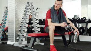 Pessoa sentada em banco de academia segura uma squeeze vermelha, destaque entre os equipamentos ao fundo. Ideal como brindes fitness, a garrafa representa hidratação e estilo durante o treino, reforçando inclusão e determinação no ambiente esportivo moderno.