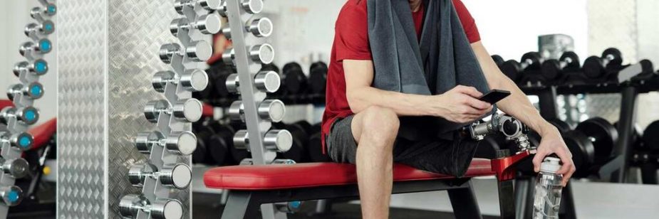 Pessoa sentada em banco de academia segura uma squeeze vermelha, destaque entre os equipamentos ao fundo. Ideal como brindes fitness, a garrafa representa hidratação e estilo durante o treino, reforçando inclusão e determinação no ambiente esportivo moderno.