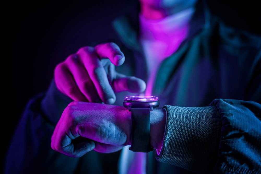 Pessoa ajustando um smartwatch com iluminação neon colorida, demonstrando tecnologia wearable moderna em um ambiente escuro.